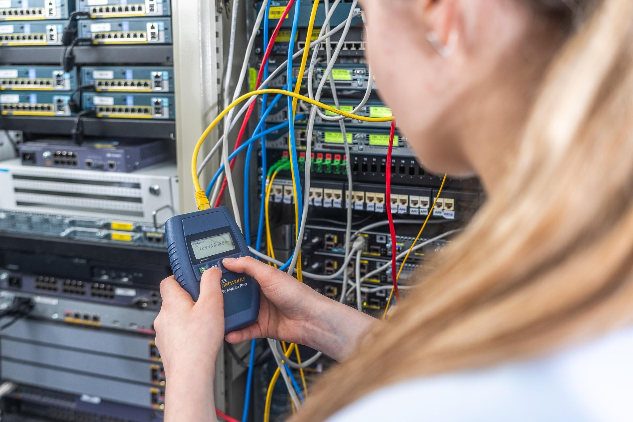 Researcher measuring a fibre connection beside network equipment in ComTech Lab.